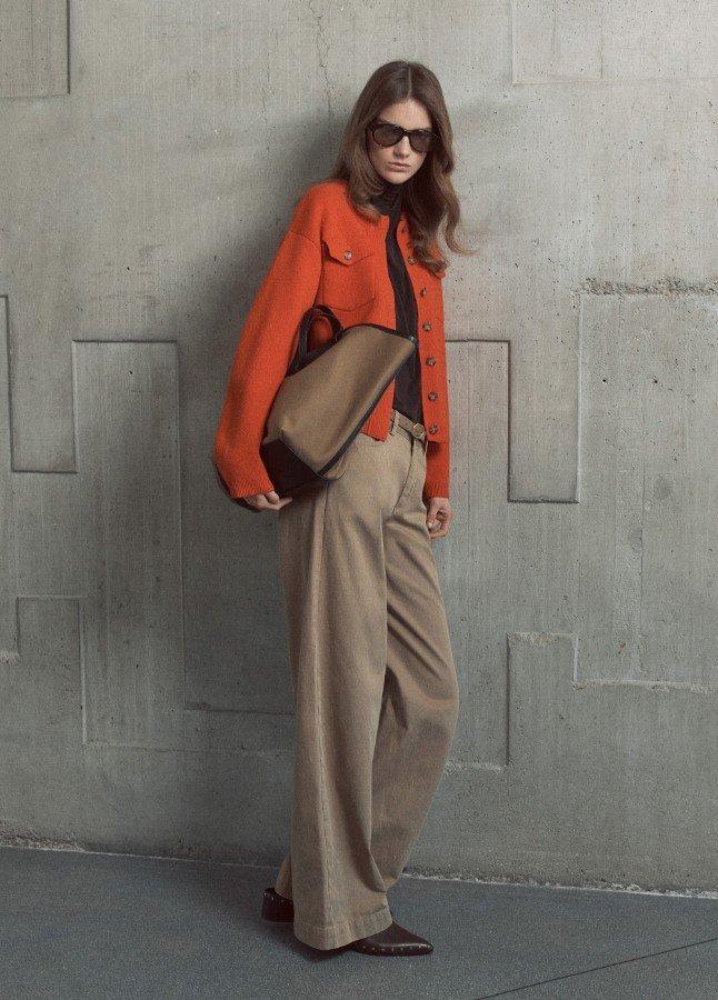 Vrouw in oranje vest, beige wijde pantalon en bruine handtas tegen grijze betonnen muur.
